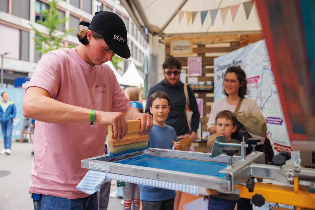 Junger Mann druckt am Stand ein Shirt, Kinder schauen zu